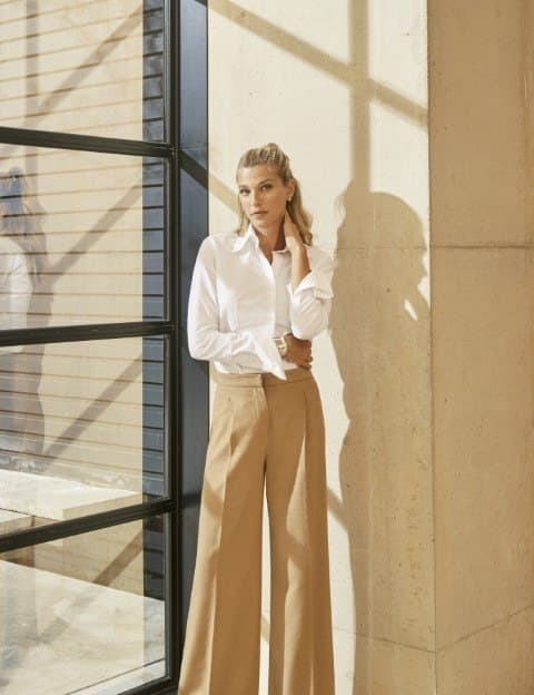 Nicole Junkermann in a white shirt and wide tan trousers, standing in bright daylight beside a glazed wall