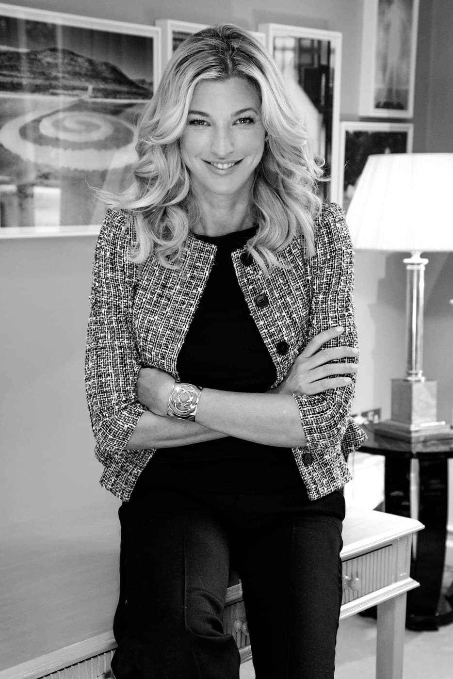 Nicole Junkermann with her arms folded in a textured jacket, a black and white portrait in a room hung with framed pictures
